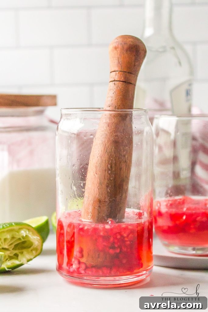 A cocktail muddler crushing raspberries, lime juice, and mint simple syrup at the bottom of a glass, releasing vibrant colors and aromas.