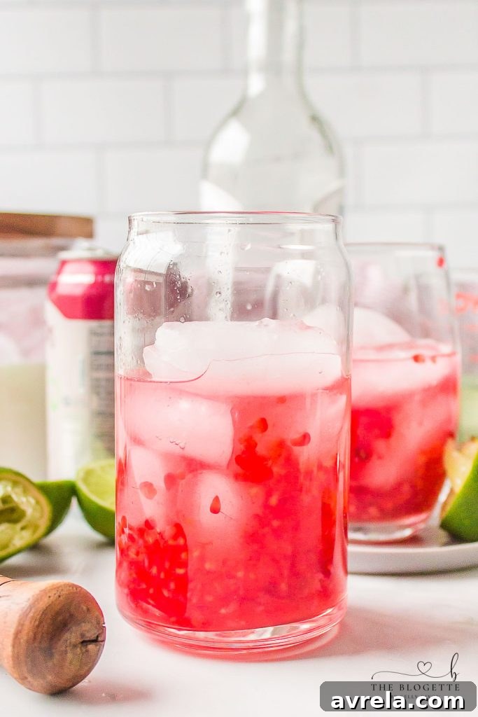 A close-up view inside a mojito glass showing muddled lime juice, raspberries, and mint simple syrup, ready for rum and sparkling water.