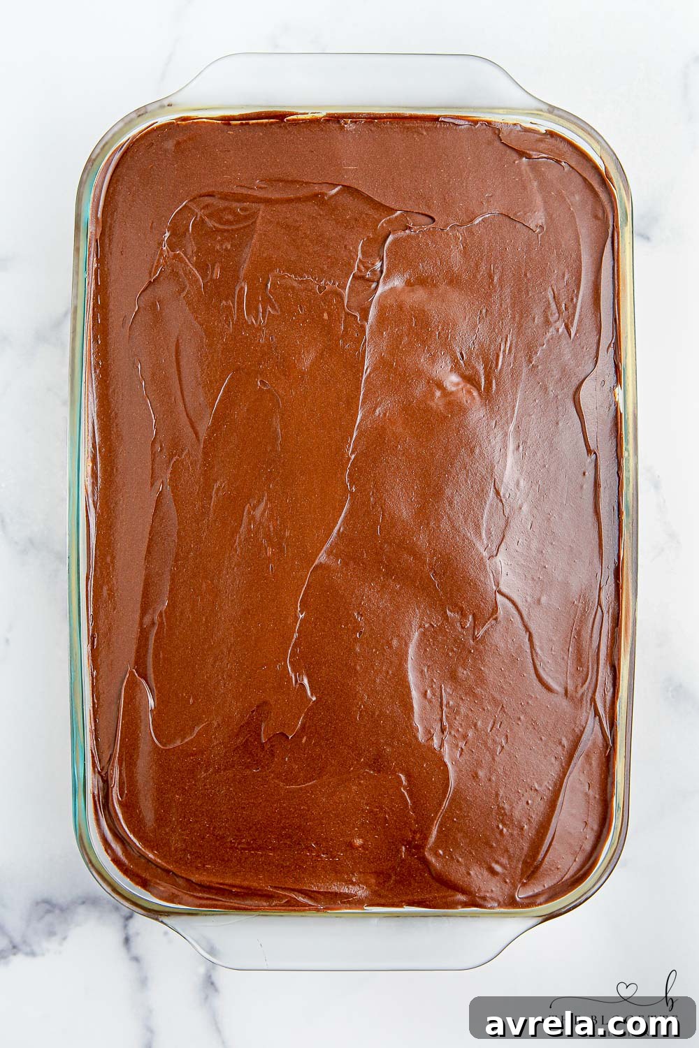 A top-down view of the Boston Cream Poke Cake, already covered with a generous, smooth layer of melted chocolate frosting, now chilling to set for perfect serving.
