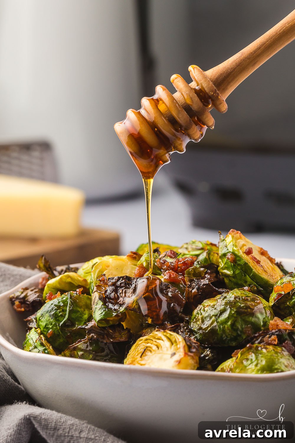 A serving bowl of Honey Bacon Brussels Sprouts being drizzled with honey.