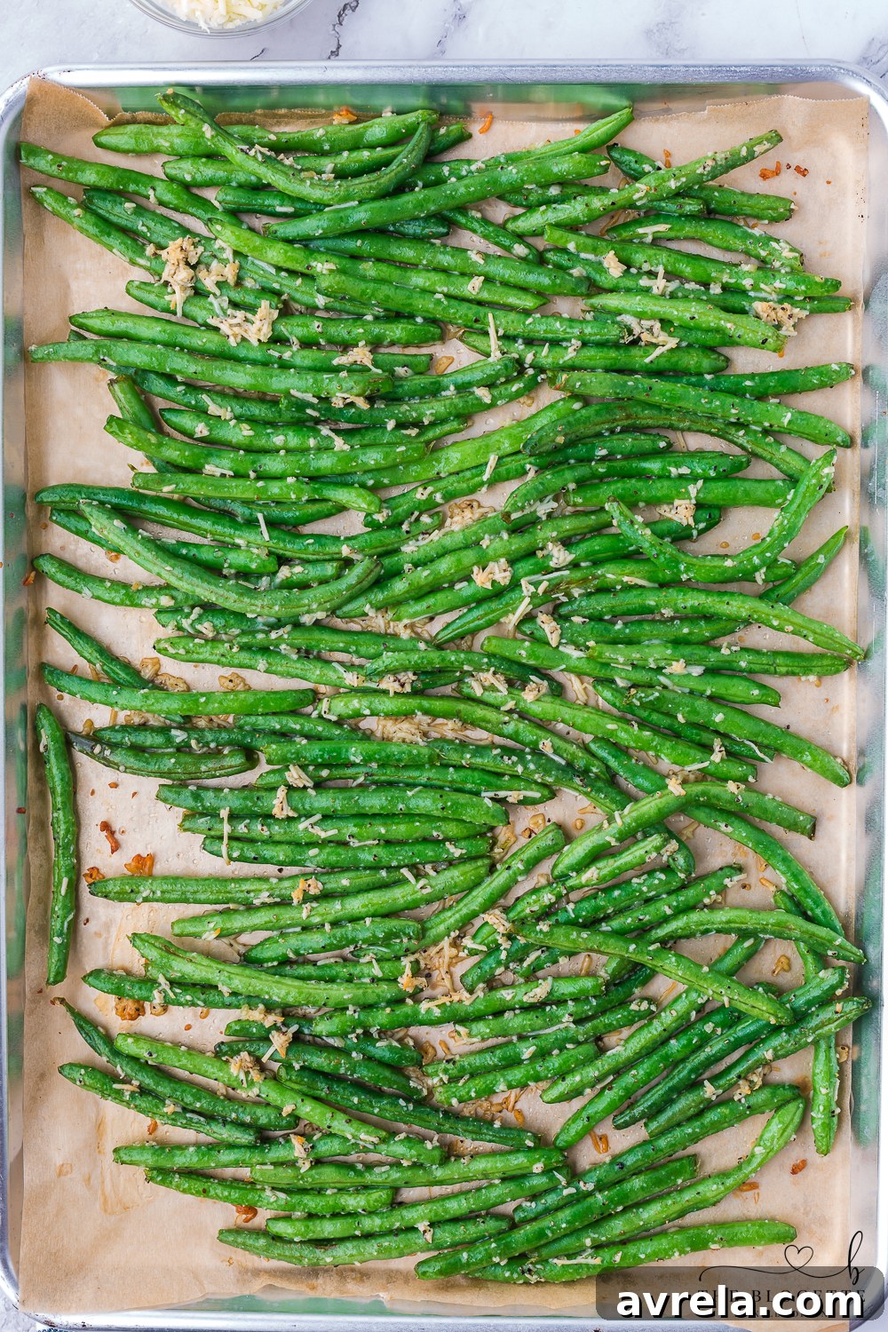 Seasoned green beans spread evenly in a single layer on a parchment-lined baking sheet, prepared for roasting in the oven.