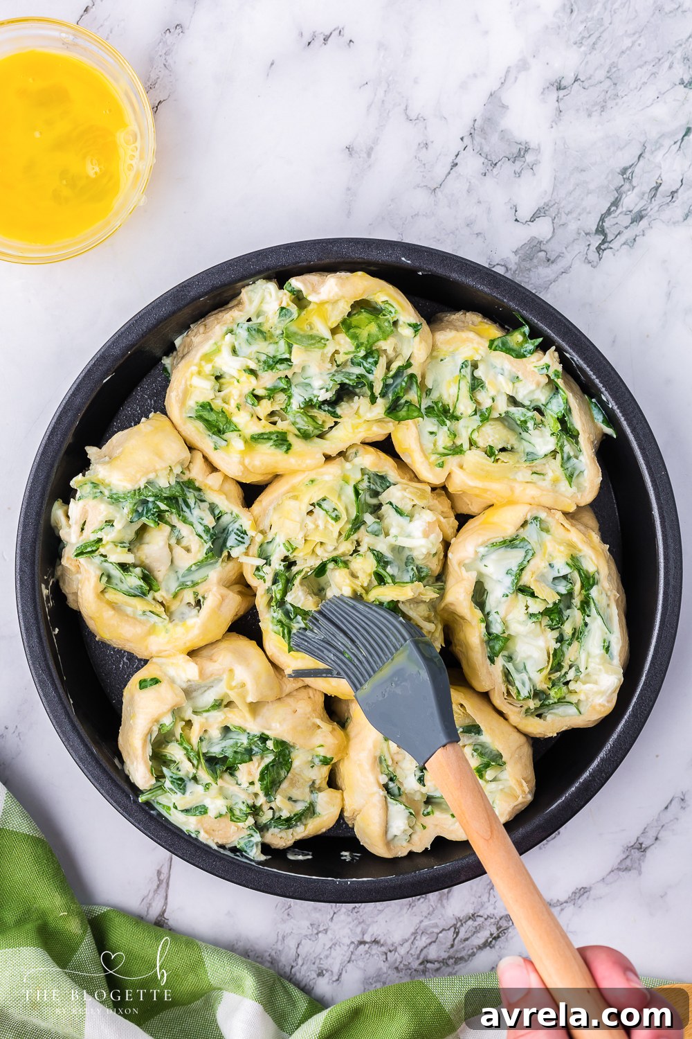 Applying an even layer of egg wash to the tops of the spinach artichoke pinwheels using a pastry brush, enhancing their golden potential.