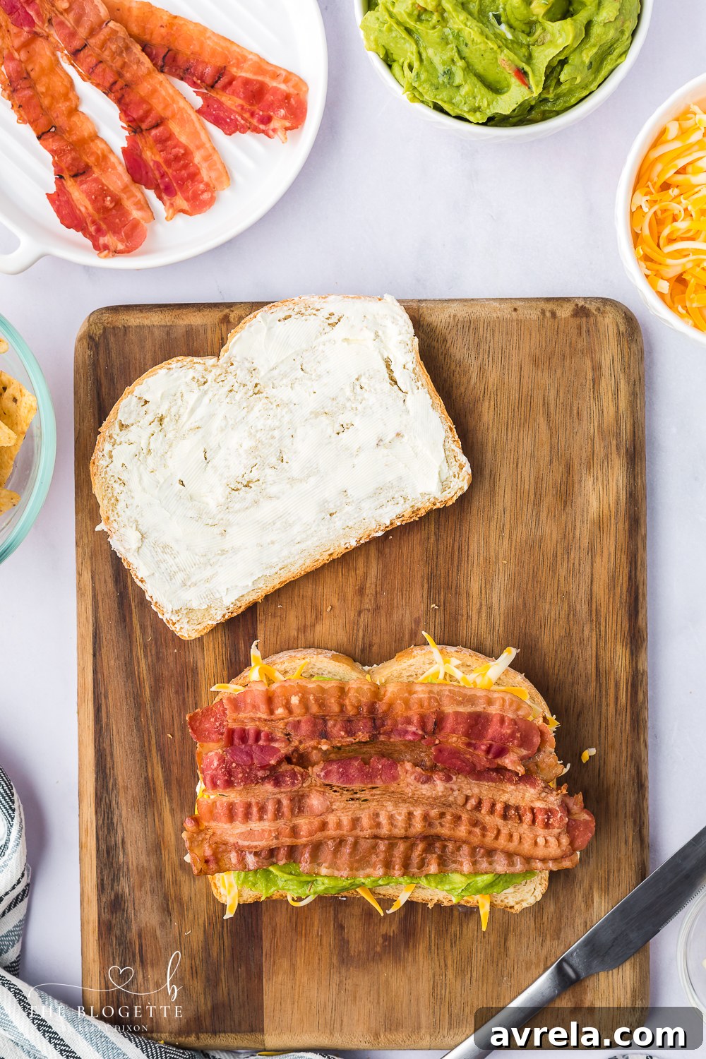 Adding crispy bacon slices over guacamole and cheese on a grilled cheese sandwich in preparation.