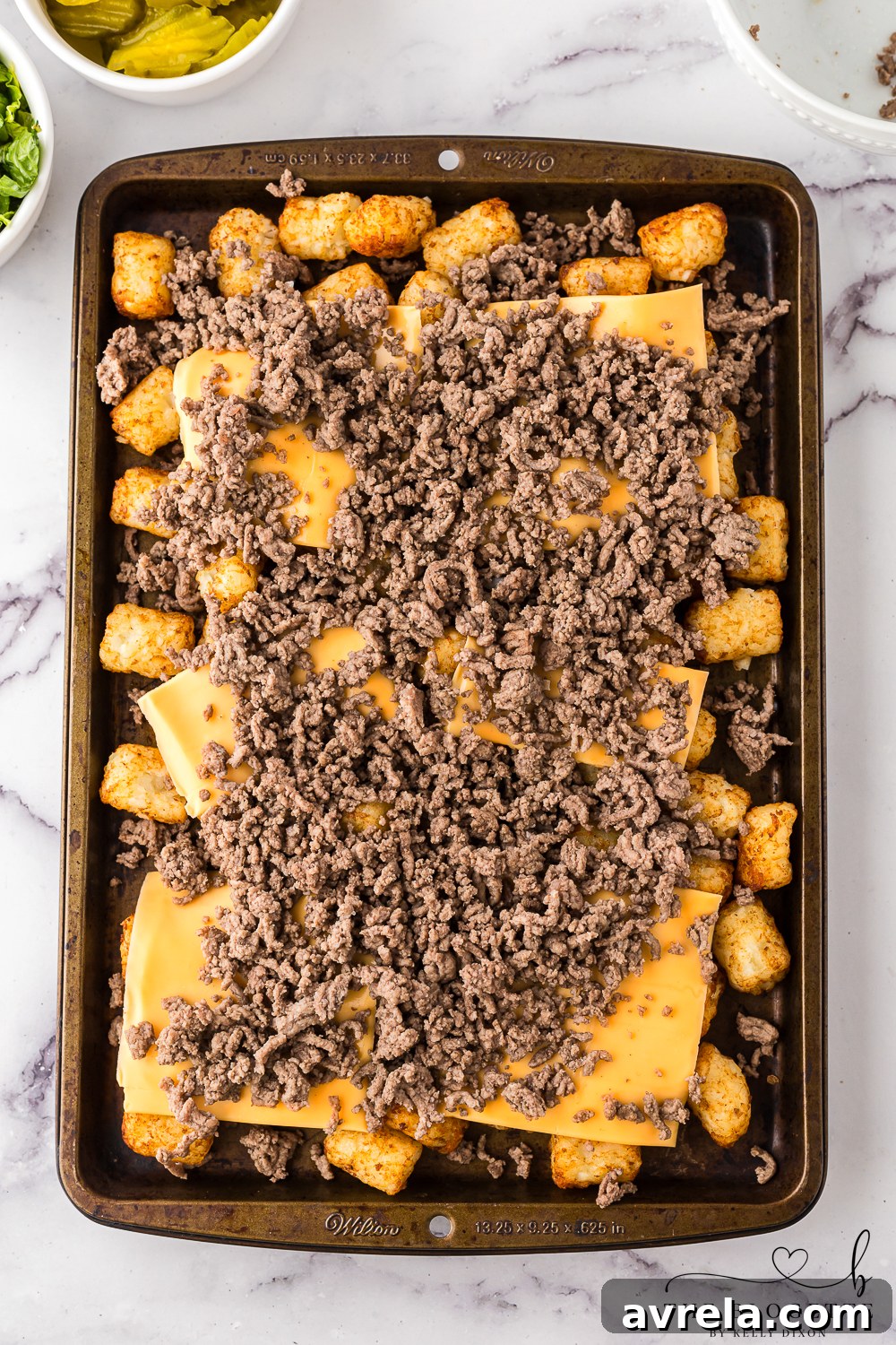 Cooked ground beef being layered over crispy tater tots and American cheese slices on a baking sheet.