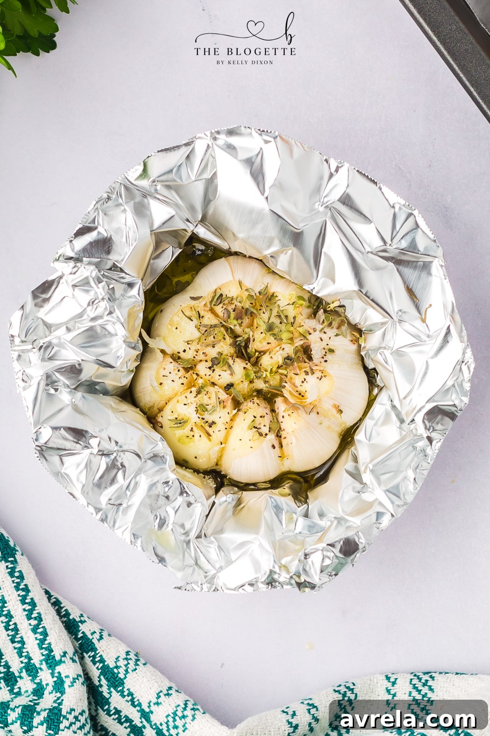A whole garlic bulb, sliced at the top and drizzled with olive oil and spices, wrapped in aluminum foil, illustrating the process of preparing garlic for roasting.