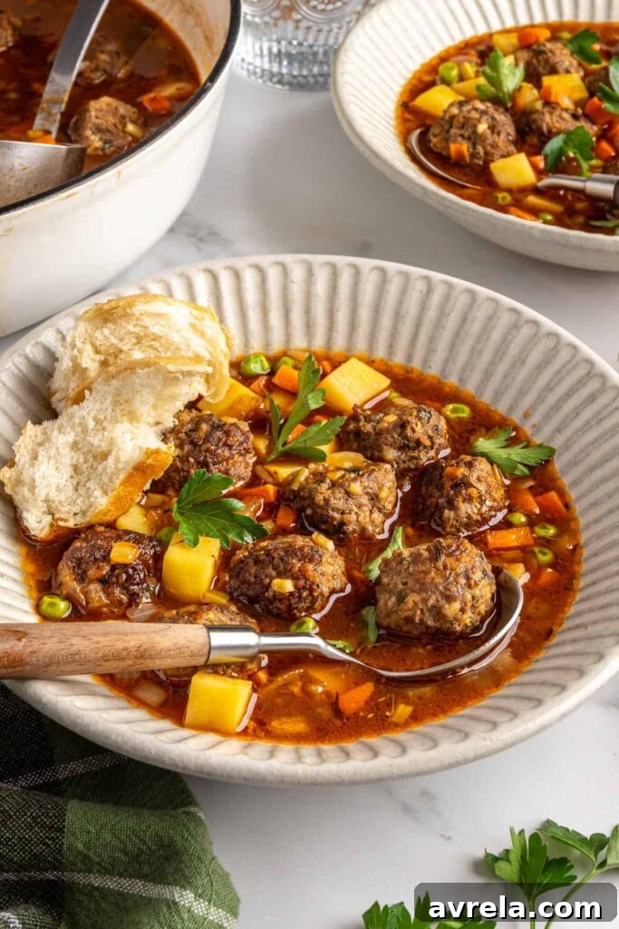 Close-up angled shot of beef meatball soup in a bowl showing meatballs, vegetables, and broth, with a piece of a baguette on the side