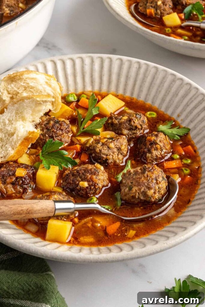 Close-up angled shot of beef meatball soup in a bowl showing meatballs, vegetables, and broth, with a piece of a baguette on the side