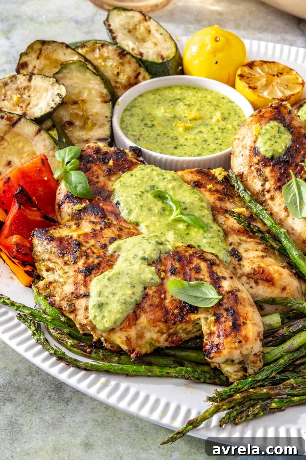 Lemon pesto grilled chicken served with grilled zucchini, asparagus, and bell peppers on a white serving platter, topped with homemade pesto and fresh lemon